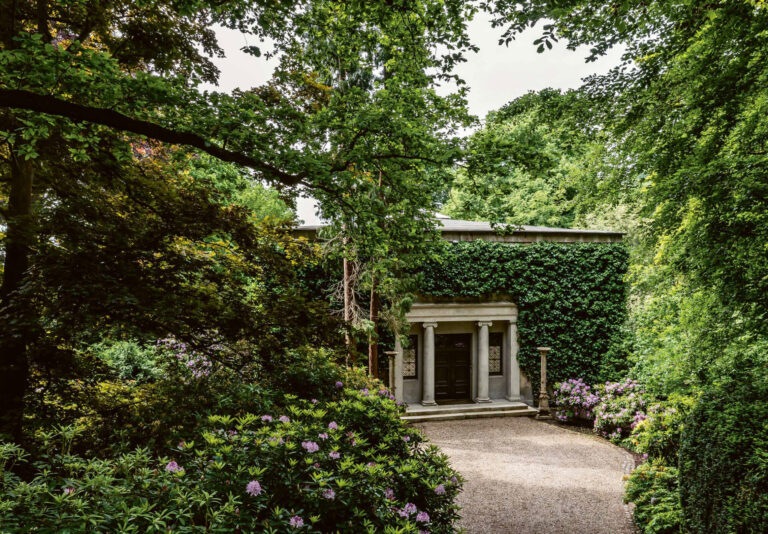 Kraftvolle Eiben und Rhododendren begleiten die mit Pflastersteinen gerahmte und mit Kies belegte Auffahrt. Die Flügeltür der Villa und die beiden Fenster aus patiniertem Messing sind in Dunkelgrün gehalten und leicht zurückversetzt. Zwei Säulen eskortieren den Blick.