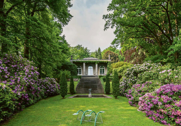 Blühende Rhododendren in Rosa- und Lilatönen säumen den Rasen, während hohe Bäume die Szene umrahmen. Brunnen und Skulptur setzen feine Akzente und verleihen dem Garten im sanften Tageslicht eine stille, märchenhafte Eleganz.