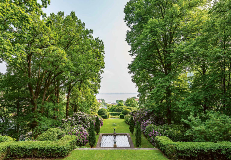 Im Vordergrund sieht man akkurat geschnittene Hecken und einen rechteckigen Brunnen. Dahinter erstreckt sich eine Rasenfläche, gesäumt von blühenden Rhododendren. Im Hintergrund öffnet sich der Blick auf ein Gewässer, das in leichtem Dunst liegt. Das Sonnenlicht wirkt warm und sommerlich.